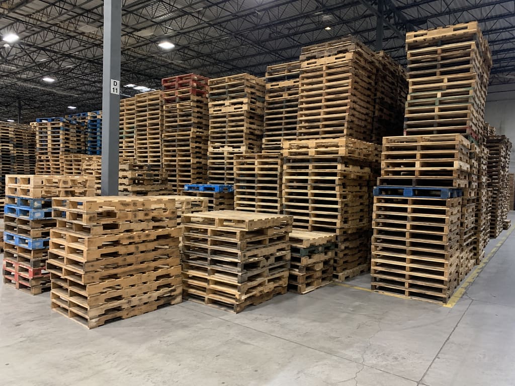 Large indoor sorting area filled with stacks of used wood pallets of varying sizes and conditions, including some blue and red pool pallets.